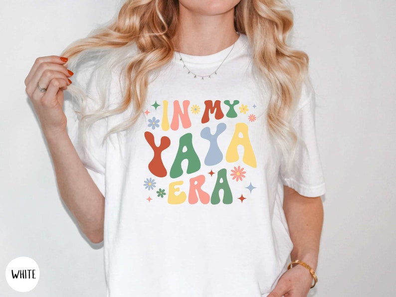 Puede incluir: Camiseta blanca con un gr&aacute;fico colorido que dice "In My Yaya Era" con flores y estrellas.