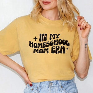 May include: A mustard yellow t-shirt with the text "In My Homeschool Mom Era" in black retro-style lettering.