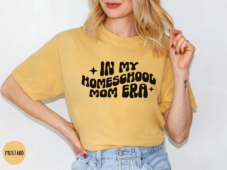 May include: A mustard yellow t-shirt with the text "In My Homeschool Mom Era" in black retro-style lettering.