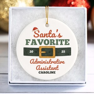 May include: A white ceramic Christmas ornament with the text "Santa's Favorite Administrative Assistant" in red and green. The ornament features a Santa hat and a belt design with the name "CAROLINE" at the bottom.