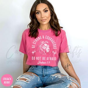 Pu&ograve; includere: Maglietta rosa con una grafica bianca di una testa di leone e il testo "Be Strong & Courageous, Do Not Be Afraid, Joshua 1:9".