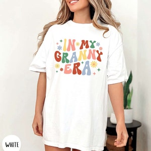 May include: White oversized t-shirt with the text "In My Granny Era" in colorful retro font with flowers and stars.