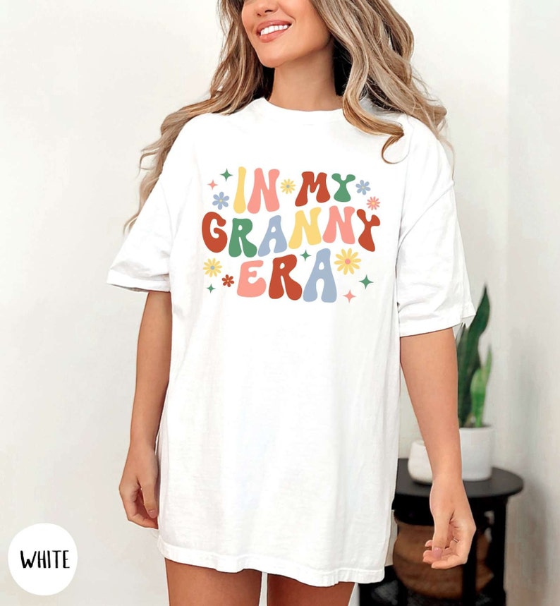 May include: White oversized t-shirt with the text "In My Granny Era" in colorful retro font with flowers and stars.