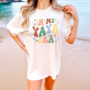Puede incluir: Una camiseta blanca con el texto "In My Yaya Era" en letras coloridas de estilo retro. El texto est&aacute; rodeado de peque&ntilde;as flores y estrellas de colores. La camiseta la lleva puesta una persona en una playa.
