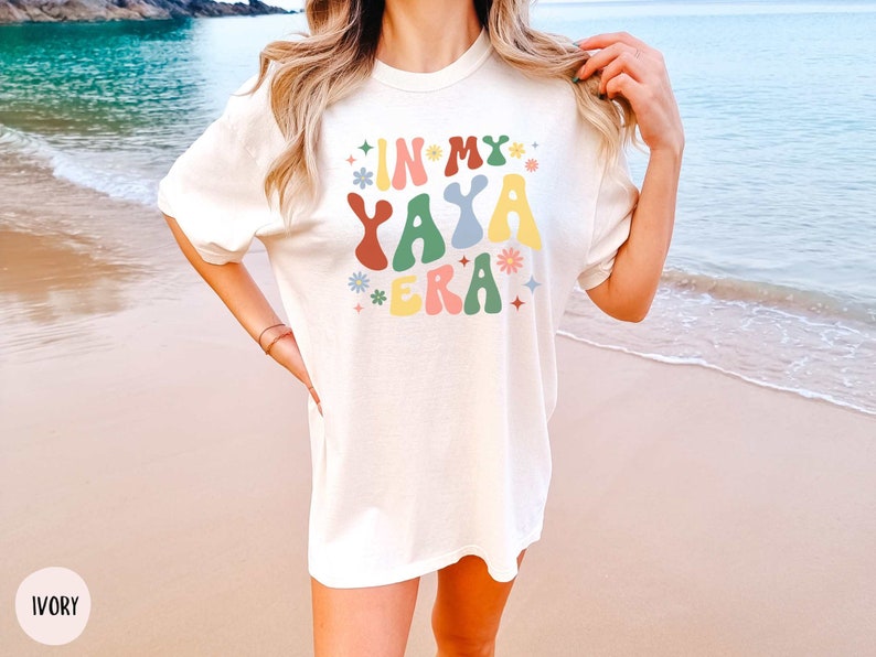 Puede incluir: Una camiseta blanca con el texto "In My Yaya Era" en letras coloridas de estilo retro. El texto est&aacute; rodeado de peque&ntilde;as flores y estrellas de colores. La camiseta la lleva puesta una persona en una playa.