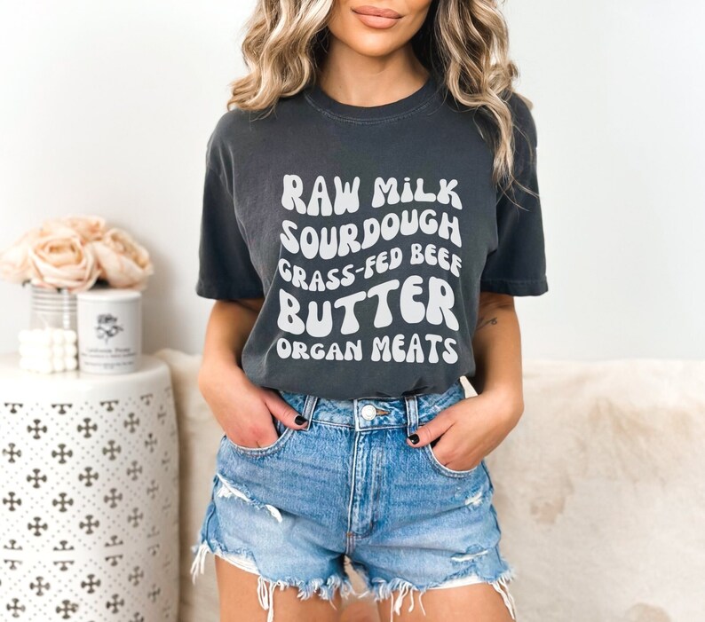May include: A dark gray t-shirt with white text that reads "Raw Milk Sourdough Grass-Fed Beef Butter Organ Meats". The person wearing the shirt is also wearing denim shorts.