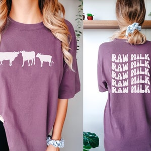 May include: A purple t-shirt with a graphic of three cows on the front and the text "RAW MILK" repeated five times on the back.