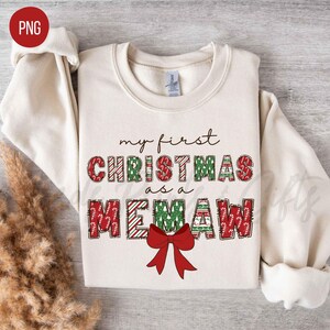 May include: Cream-colored sweatshirt with the text "my first CHRISTMAS as a MEMAW" in green and red. The word "CHRISTMAS" is in a festive pattern, and a red bow is at the bottom.