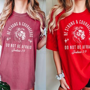 Pu&ograve; includere: Due t-shirt da donna con una grafica di un leone e il testo "Be Strong & Courageous Do Not Be Afraid Joshua 1:9". La maglietta a sinistra &egrave; rosso mattone e la maglietta a destra &egrave; rossa.