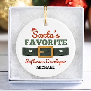 May include: A white, round Christmas ornament with the text "Santa's FAVORITE Software Developer MICHAEL". The design includes a Santa hat, a green belt with a gold buckle, and the years 20 and 25.