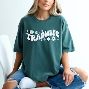 Puede incluir: Una camiseta azul turquesa con el texto "TRADWIFE" en blanco, con una fuente retro y detalles de flores.