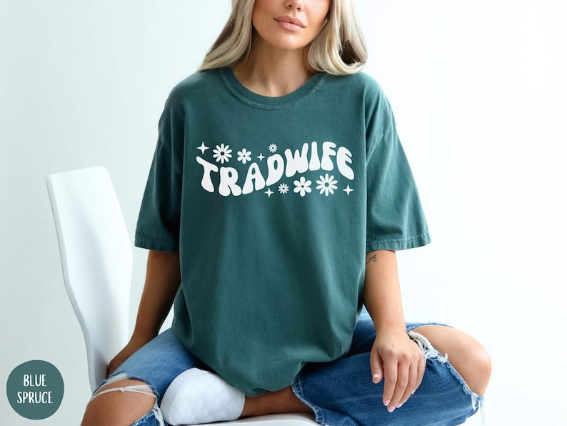 Puede incluir: Una camiseta azul turquesa con el texto "TRADWIFE" en blanco, con una fuente retro y detalles de flores.