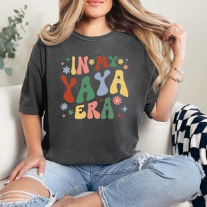 Puede incluir: Una camiseta gris oscuro con el texto "In My Yaya Era" en letras coloridas de estilo retro. La camiseta tiene un corte holgado y est&aacute; hecha de una tela suave y c&oacute;moda.