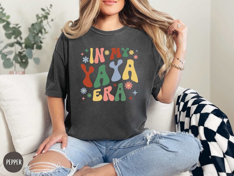 Puede incluir: Una camiseta gris oscuro con el texto "In My Yaya Era" en letras coloridas de estilo retro. La camiseta tiene un corte holgado y est&aacute; hecha de una tela suave y c&oacute;moda.