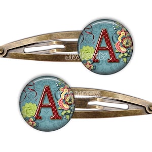 May include: A pair of bronze hair clips with a blue and red floral design and the letter A in the center.