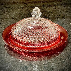 May include: A clear glass butter dish with a diamond pattern on the lid and a red base. The lid has a decorative finial. The dish is designed to hold butter and is a classic kitchen item.