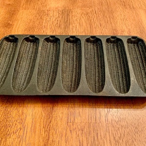 May include: A cast iron cornbread pan with eight individual molds shaped like corn cobs. The pan is black and has a slightly worn appearance.
