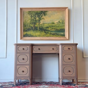 May include: A wooden desk with a light brown finish featuring six side drawers and one central drawer, each with a small round knob. Above the desk hangs a rectangular landscape painting in a wooden frame depicting trees, a field, and a pond.