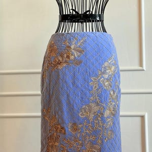 May include: A light blue skirt with a quilted pattern and gold floral embroidery. The skirt is displayed on a black wire dress form. The floral design is concentrated on the lower half of the skirt.