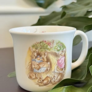May include: White ceramic mug featuring a charming illustration of rabbits in a garden setting. The artwork depicts several bunnies in various poses, with soft pastel colors. The mug has a curved handle and a classic shape, suitable for hot beverages.