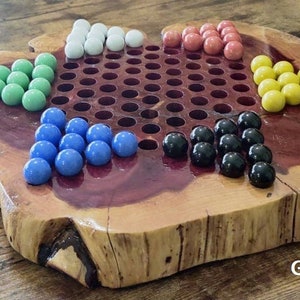 May include: A wooden game board with a red stained center and a natural wood finish. The board has a grid pattern with 121 holes and is surrounded by a raised edge. There are 10 marbles in each of five colors: white, green, blue, black, and yellow. The board is labeled "Gloss Finish".