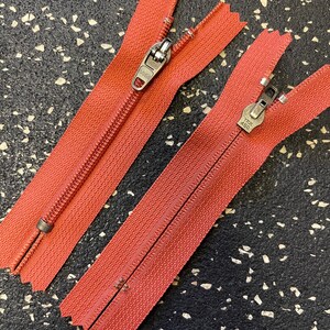 May include: Two red zipper pulls with metal sliders. The zippers are separated and laid out on a black and white speckled surface.