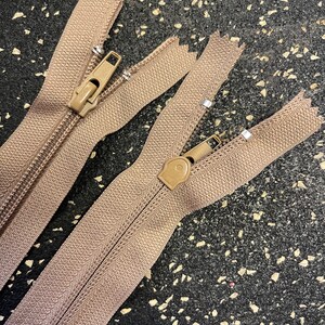 May include: Two brown zippers with metal sliders. The zippers are laid out on a black surface.