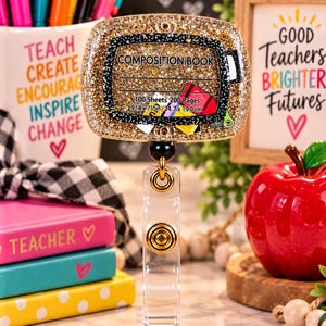 May include: A rectangular badge reel featuring a gold glitter-filled acrylic topper shaped like a composition book. The topper displays the text COMPOSITION BOOK, 100 Sheets 200 Pages, and 9 3/4 x 7 1/2 in / 24.7 x 19.0 cm. It has a clear plastic strap and a gold metal snap button.