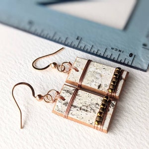 May include: Close-up of handmade earrings. Each earring features a rectangular, birch bark-like design with copper wire accents and gold-colored beads. The ear hooks are a matching copper color. A ruler is in the background.