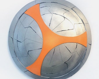 Puede incluir: Un escudo plateado con una superficie agrietada y un diseño de estrella naranja de cuatro puntas.