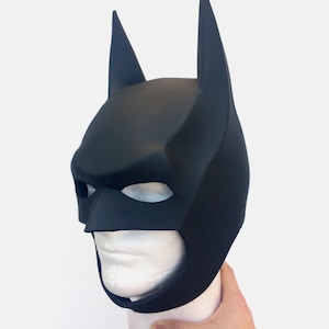 Peut inclure: Un masque noir de Batman avec des oreilles pointues et des ouvertures pour les yeux. Le masque est en matériau mat et est présenté sur une tête de mannequin blanche. Accessoire de costume.