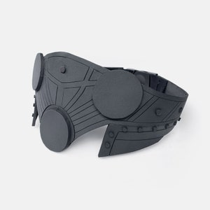 May include: A dark gray, stylized mask with circular and geometric design elements. The mask features multiple circular accents and a series of rivets along the edges. The overall design suggests a futuristic or steampunk aesthetic.