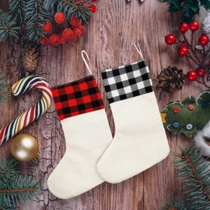 May include: Two Christmas stockings with white bodies and buffalo check tops in red and black, and black and white. The stockings are on a wooden surface with festive decorations, including a candy cane, pine needles, and berries.