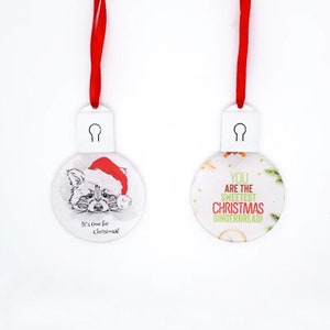 May include: Two round white ornaments with red ribbon hangers. One features a raccoon wearing a Santa hat and the text It's time for Christmas! The other displays the text YOU ARE THE SWEETEST CHRISTMAS GINGERBREAD! with small pine and orange accents.
