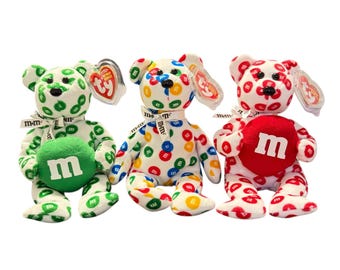May include: Three Ty Beanie Baby bears with M&M's candy designs. One bear is green with white polka dots and a green M&M's candy. Another is white with colorful M&M's candies. The third is red with white polka dots and a red M&M's candy.