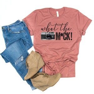 May include: A dusty rose t-shirt with the text "what the m*ck!" and a vintage camera graphic. The shirt is paired with blue jeans and brown ankle boots.