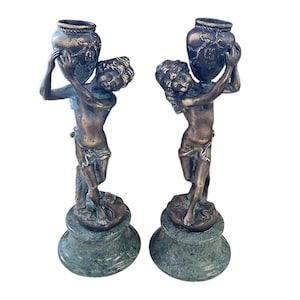 May include: A pair of bronze-toned sculptures depicting figures holding ornate urns. The figures stand atop green marble bases. The urns feature detailed floral designs. The sculptures are likely decorative items.