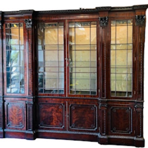 May include: A large, antique, mahogany bookcase with glass doors and intricate carvings. The bookcase has three sections, each with two doors and multiple shelves. The doors are framed with a decorative molding and have a grid pattern of glass panes.