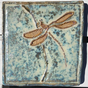 May include: Square ceramic tile with a dragonfly design. The tile has a textured, mottled blue and cream background. A raised dragonfly in shades of brown and tan is centered on the tile, with delicate lines suggesting its wings and body.