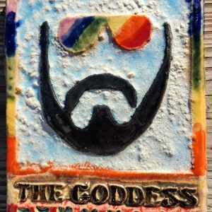 May include: A colorful ceramic tile with a cartoon face wearing rainbow sunglasses and a black beard. The tile has a white background and the words "THE GODDESS" are written in gold letters at the bottom. The tile is decorated with red hearts along the bottom edge.