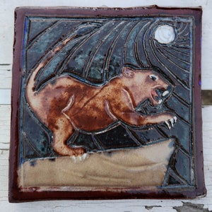 May include: A ceramic tile with a brown and white image of a snarling animal with a moon in the background.