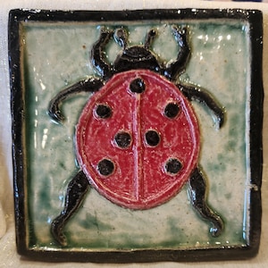 May include: Square ceramic tile featuring a red ladybug with black spots and legs. The ladybug is centered on a light green background, framed by a black border. The tile has a glossy finish, suggesting it's handmade.