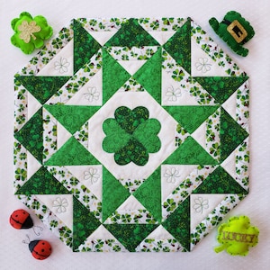 May include: A green and white quilted table runner with a shamrock design. The runner is surrounded by four felt shamrocks and a felt leprechaun hat.
