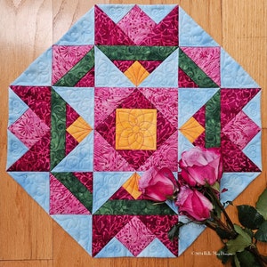 May include: A quilted table runner with a geometric pattern in shades of pink, blue, green, and yellow. The runner is decorated with three pink roses.