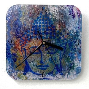 May include: Square wall clock with a colorful abstract design featuring a serene Buddha face. The clock face has blue, orange, and white hues, with black clock hands and numerals at 3, 6, 9, and 12.