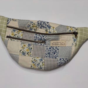 May include: A quilted fanny pack with a patchwork design. The bag features a mix of floral, gingham, and solid fabric squares in shades of blue, gray, and cream. It has two zippered compartments and a light green checkered fabric on the sides. The text "HANDMADE BY The Modern Doe Co." is visible.