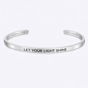 Puede incluir: Una pulsera de plata con la inscripción grabada "LET YOUR LIGHT SHINE". La pulsera también presenta las inscripciones grabadas "Matthew 5:16" y "NCOURAGE". La pulsera tiene un diseño sencillo y elegante.