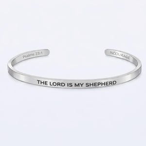 Puede incluir: Una pulsera de plata con la inscripción "THE LORD IS MY SHEPHERD" en letras negras. La pulsera también presenta el texto "Psalms 23:1" y "NCOURAGE". Una joya de temática religiosa.