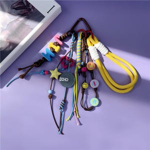 Peut inclure: Un assortiment de porte-clés et de lanières colorés. Les porte-clés présentent des perles de lettres, des breloques en forme d'étoile et des breloques rondes avec des visages souriants et le mot "ECHO". Les lanières sont jaunes et blanches.