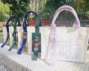 Può includere: Quattro borse tote con diversi design. Le borse sono bianche con cinturini neri e design colorati. I design includono un motivo floreale, una grafica di un personaggio dei cartoni animati, una grafica di un edificio e il testo "You Never Walk Alone" con le parole "Armyland" sotto.